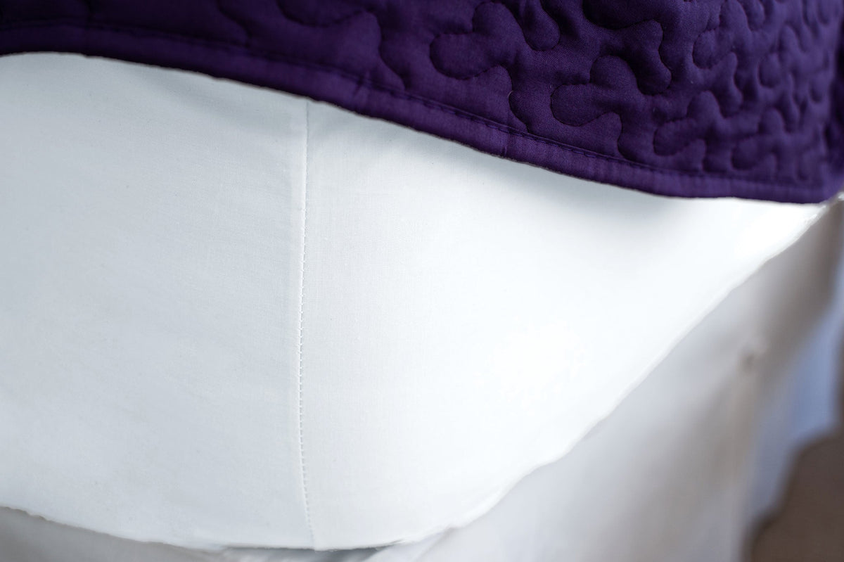 Corner of mattress made with white organic cotton fitted sheet with a purple bedspread.