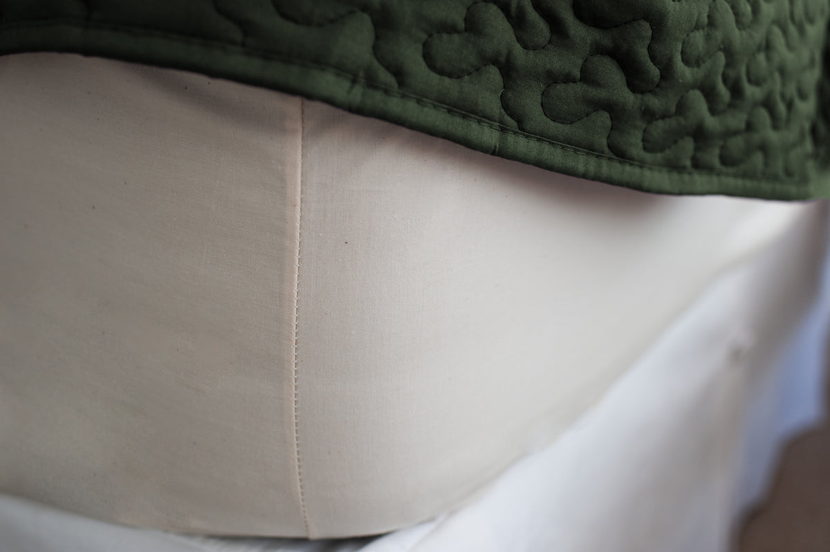 Corner of mattress made with organic cotton fitted sheet in natural color with a green bedspread.