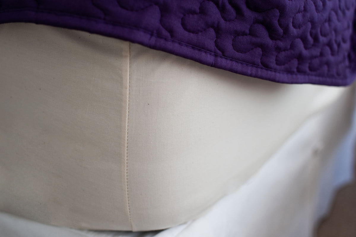 Corner of mattress made with organic cotton fitted sheet in natuCloseup of Corner Color Natural Single Fitted Bed Sheet [Separate] Cotton Made in USAral color with a purple bedspread.